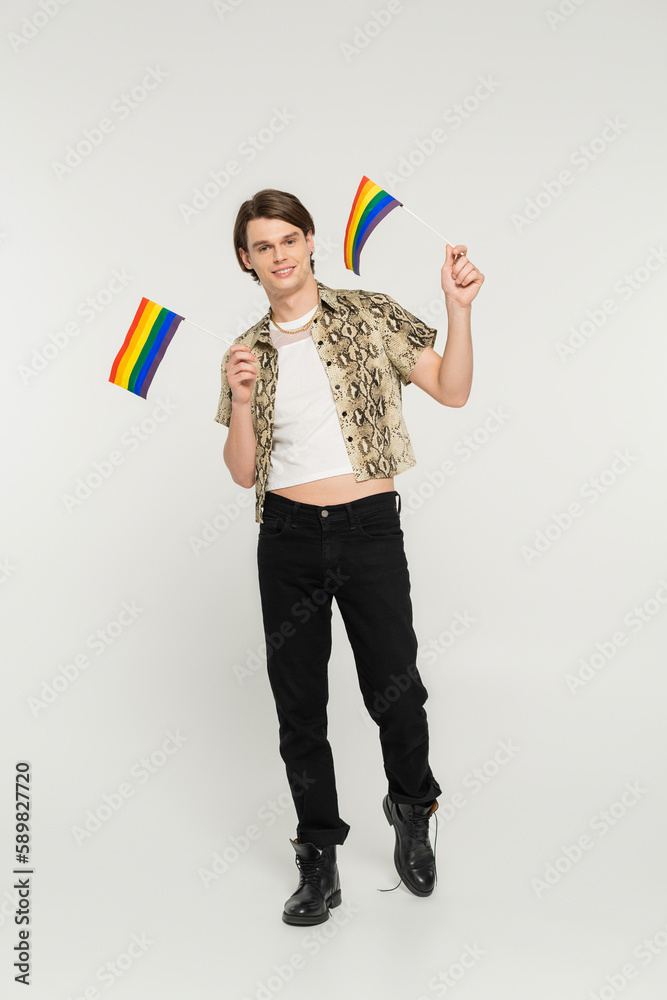 full length of positive pansexual model in black pants and snakeskin print blouse holding small lgbt flags on grey background. full length of positive pansexual model in black pants and snakeskin print blouse holding small lgbt flags on grey background.