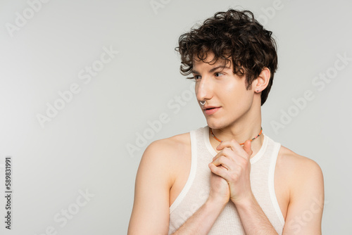young brunette bigender model with wavy hair and nose piercing looking away isolated on grey.