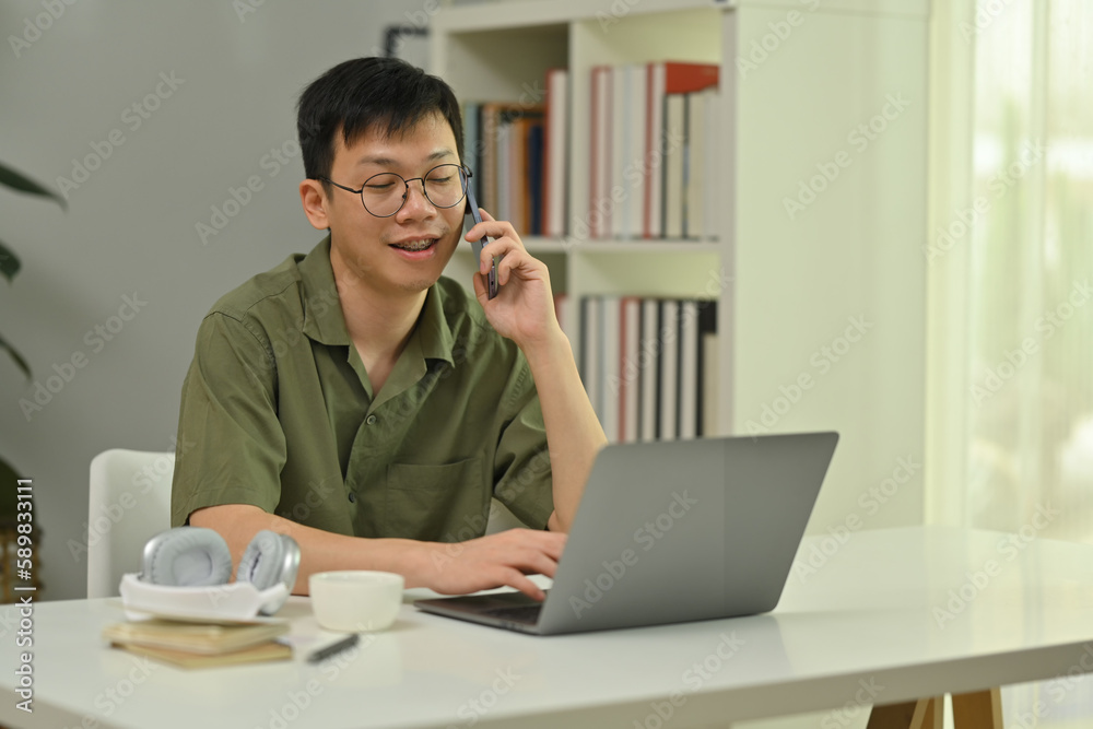 Positive millennial man in glasses talking on cellphone, sitting at working desk with laptop computer Positive millennial man in glasses talking on cellphone, sitting at working desk with laptop computer