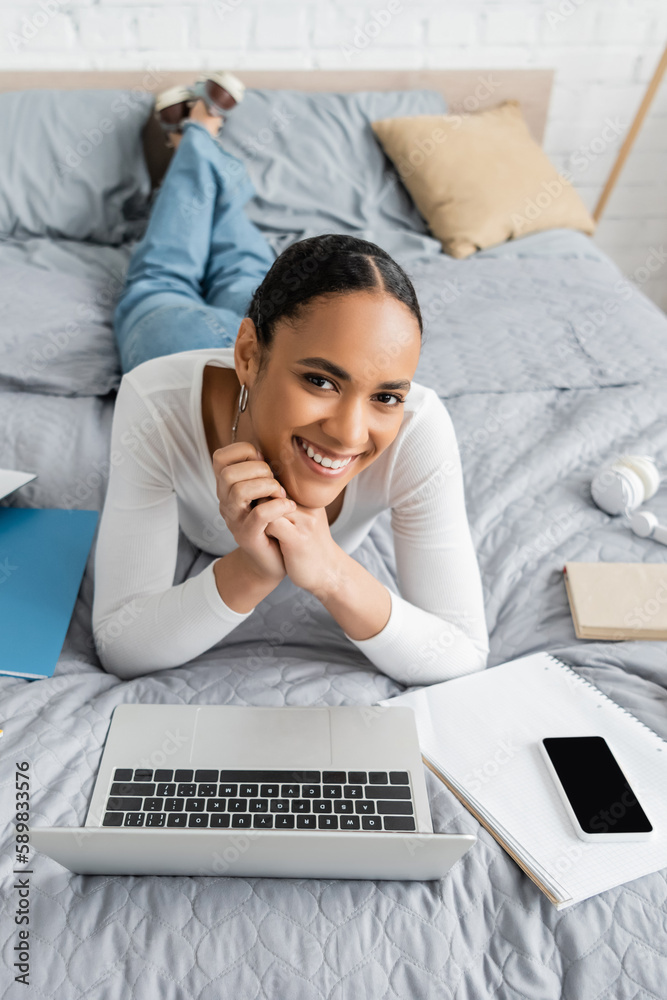 Cheerful african american student lying near gadgets and notebooks on bed. Cheerful african american student lying near gadgets and notebooks on bed.