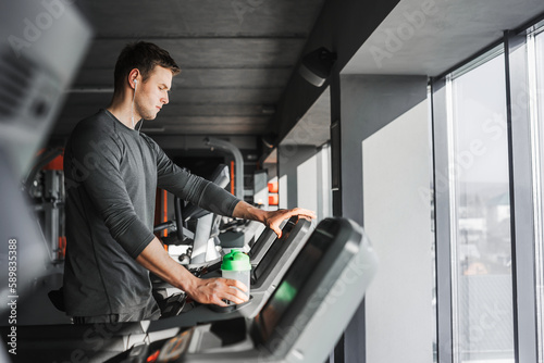 Young athletic man standing on treadmill in gym. In one hand he holds a green water bottle and the other touches the treadmill screen.