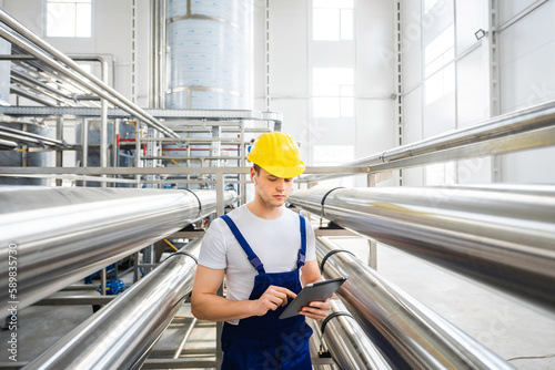 A young male specialist in a yellow hard hat uses tablet PC for controls the technological processes of equipment for industrial water purification. Industrial worker indoors in factory. photo