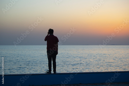 Young man watching the sea at sunset. man sitting on a wall by the ocean. Embracing the beauty and power of the ocean at sunset.