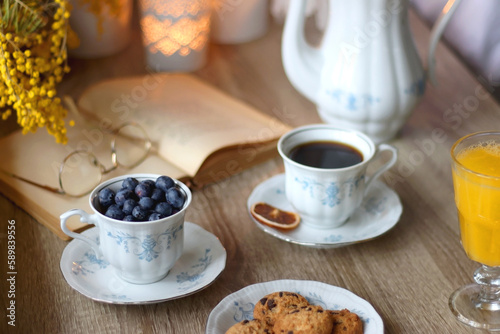 Cup of tea or coffee, plate of cookies, cup of blueberries, plate of chocolate, glass of juice, book, reading glasses, teapot, flowers and lit candles on the table. Brunch or afternoon tea concept. 