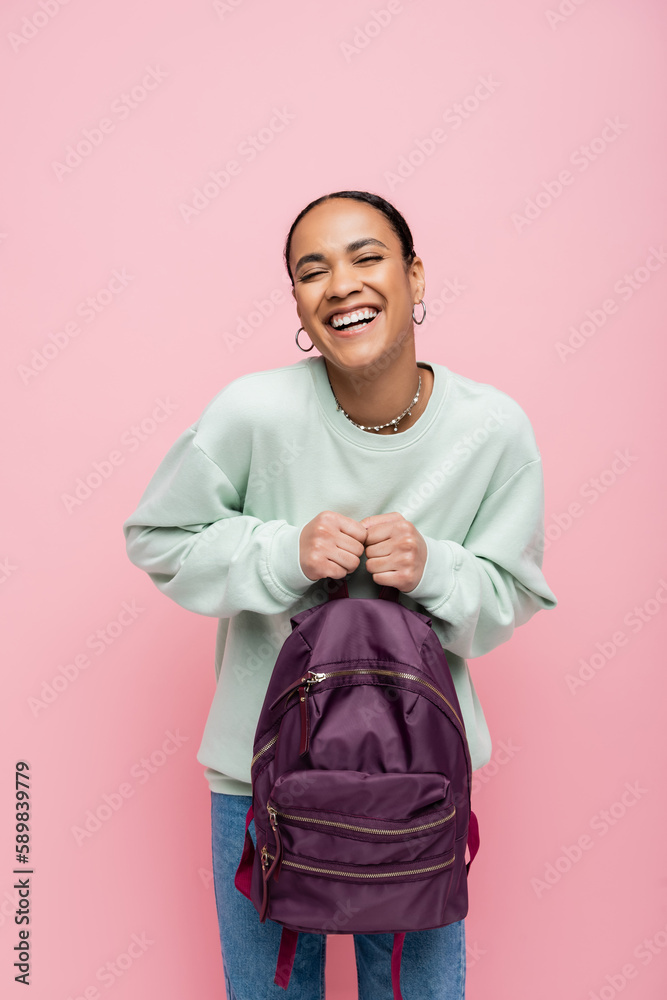 joyful african american student in sweatshirt holding purple backpack isolated on pink. joyful african american student in sweatshirt holding purple backpack isolated on pink.