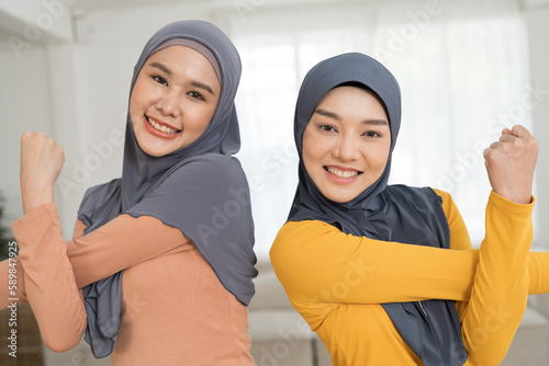 Two Asian young muslim women in hijab doing work out, stretch arm at home. Asian muslim women exercising with happy and smiling in living room photo