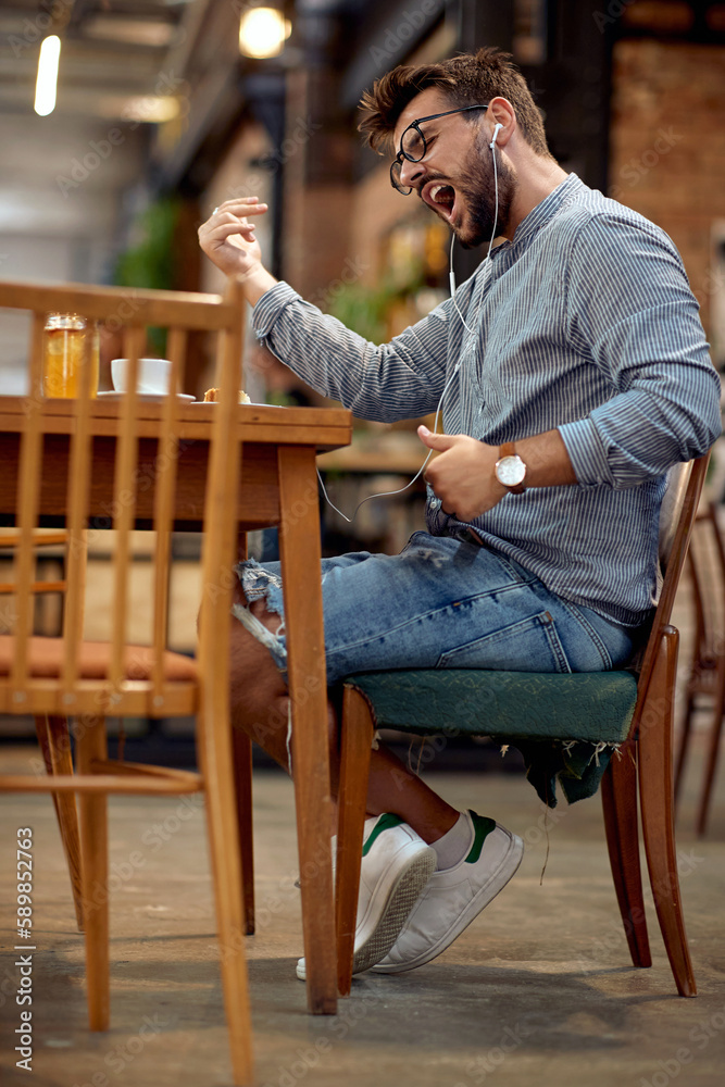 Handsome young man siting in cafe, playing music and dancing to it. Cafe background. Handsome young man siting in cafe, playing music and dancing to it. Cafe background.