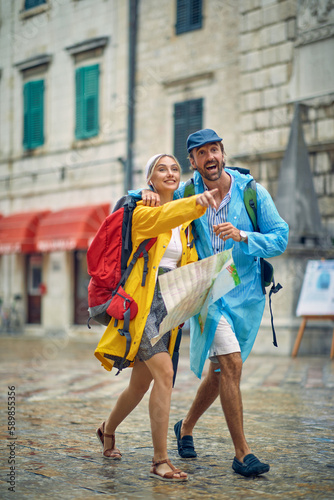 Young beautiful couple in raincoats on walk through city. Feeling amused and joyful, woman pointing forward. Travel, togetherness, city, lifestyle concept.