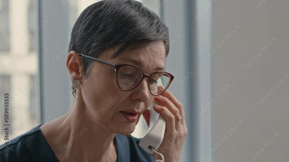 Medium closeup of confident mid adult short haired female lawyer in eyeglasses making phone call while sitting at desk in successful law firm