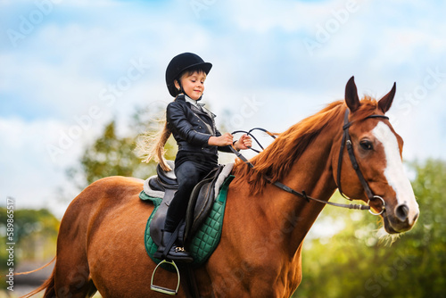 Small child in jockey outfit is riding horse on blue sky with clouds background. School of riding and equestrian sports. © Olha