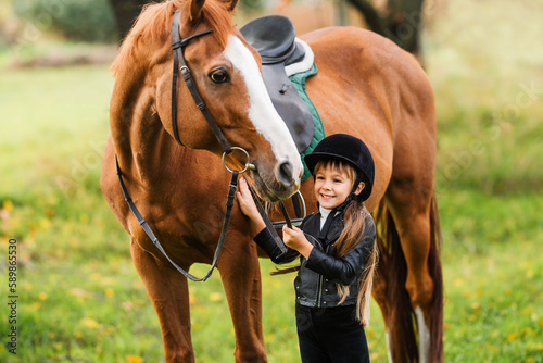 Happy child is holding horse by bridle on green meadow. Little kid in jockey outfit play with horse.