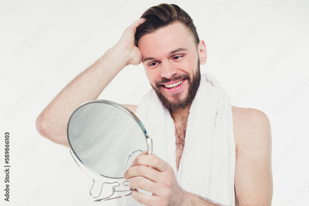 Close up portrait of a handsome bearded man with towel looking at his reflection in the mirror in bathroom isolated on white background