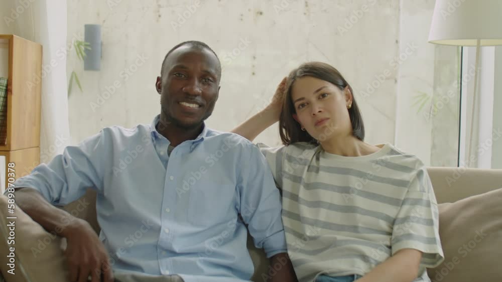 Medium shot of diverse family couple sitting together on sofa in living room, watching TV and chatting while spending leisure time together at home Medium shot of diverse family couple sitting together on sofa in living room, watching TV and chatting while spending leisure time together at home
