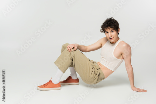 full length of brunette pansexual model in beige pants looking at camera while posing on grey background. photo