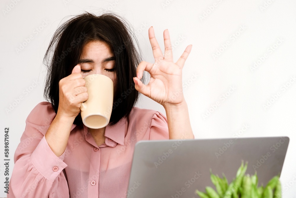 Tired Asian worker lack of sleep and energy drinking a cup of coffee and trying to work sitting at the office with a laptop on desk