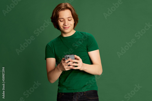 Portrait of young student, guy with long hair holding cell phone on hands and smiling with pleasure over green studio background. Online, gadgets photo