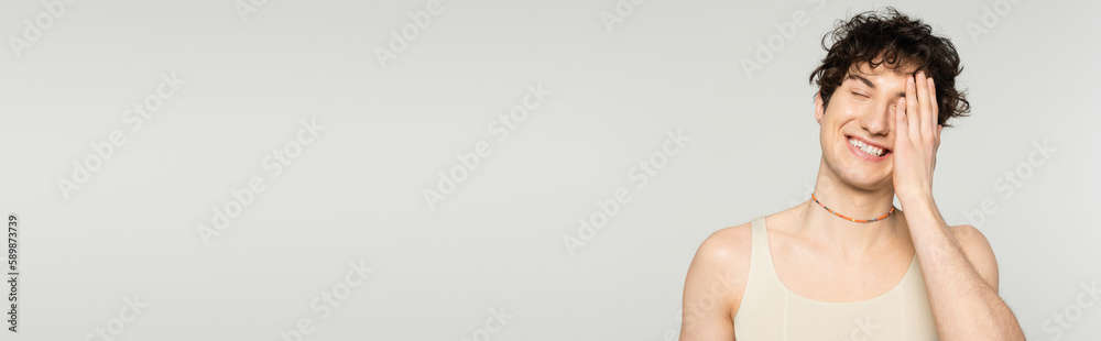 cheerful pansexual model in tank top covering face with hand and smiling with closed eyes isolated on grey, banner. cheerful pansexual model in tank top covering face with hand and smiling with closed eyes isolated on grey, banner.