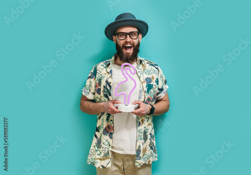 A hipster guy with a beard in a hat and a Hawaiian shirt holds a neon lamp in the shape of a flamingo in his hands. On a blue studio background