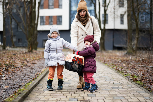 Mother and daughters walking with kitten in travel plastic cage carriage outdoor at park.