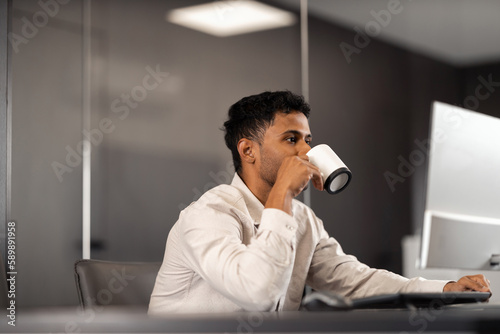 A multiethnic lawyer is drinking coffee while working on a desktop computer in an elegant office.