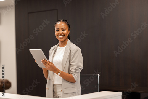 The young African-American female receptionist stands behind the reception desk with a digital tablet in her hands and smiles at the camera. photo