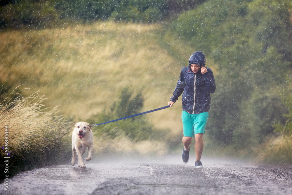 Φωτογραφία Man with dog on leash running together on wet rural road in heavy rain