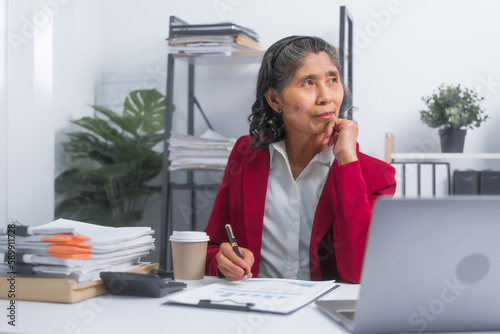 Confident asian elderly businesswoman in red suit working at desk planning bookkeeping project, thinking and reading, sitting at workplace.