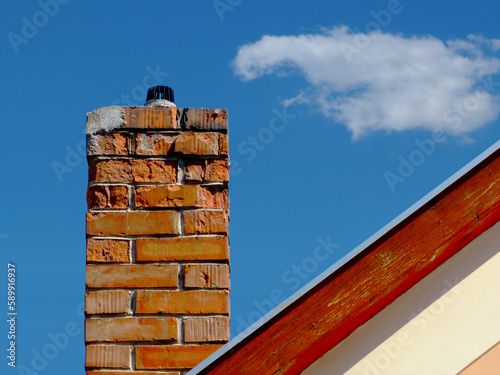 Isolated damaged clay brick chimney with weathered and spalling surface. wood trim on house gable end wall. metal flashing. white stucco exterior elevation. strong shadow. blue sky. fluffy white cloud photo