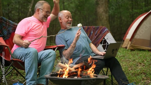 Two gay men with pride flag and tent in tent talking to laptop on video chat.