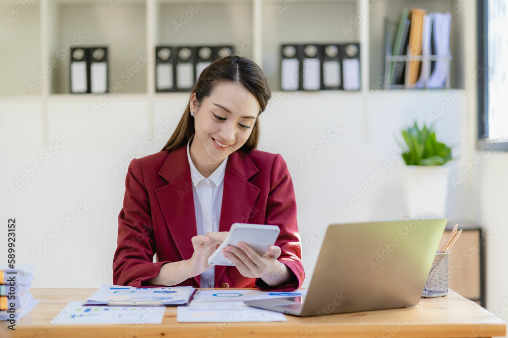 Smiling Asian businesswoman working on tablet computer at her desk in bright modern office Calculating financial report expenses with graph documents Smiling Asian businesswoman working on tablet computer at her desk in bright modern office Calculating financial report expenses with graph documents