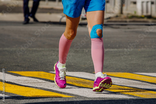 legs female runner in pink compression socks running pedestrian crossing on road, knee in kinesiotaping injury protection photo