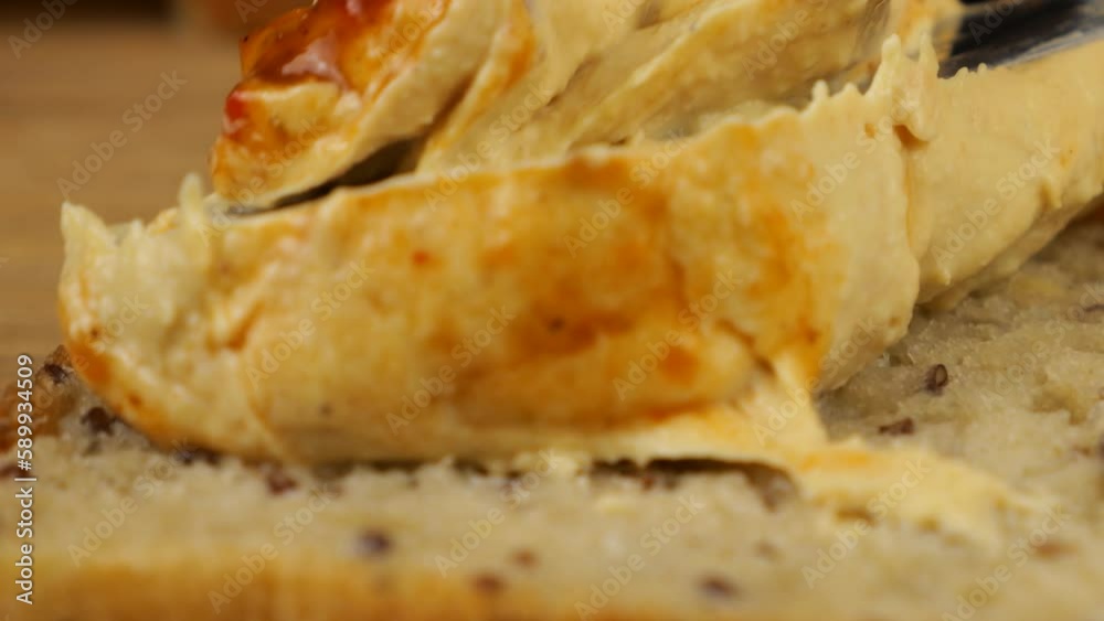 Hummus is spread with a knife on a piece of multigrain bread on a wooden cutting board. Food for vegans. Hummus is spread with a knife on a piece of multigrain bread on a wooden cutting board. Food for vegans.