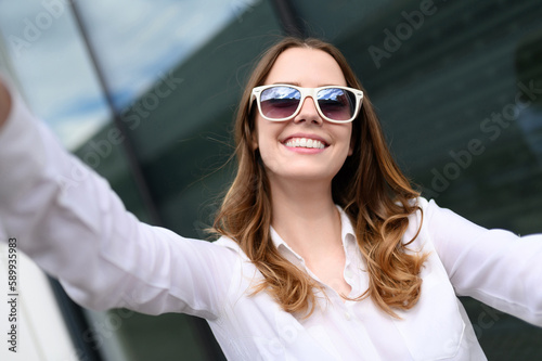 Young happy woman in summer clothes, sunglasses posing on the background of the business center, making a selfie portrait. Summer, technology concept