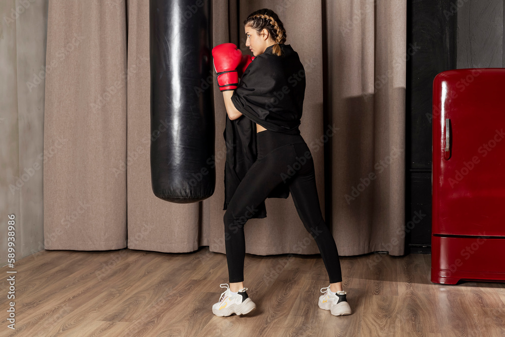 A girl in red boxing gloves, black tight leggins and a towel on her shoulders is training with a punching bag.
