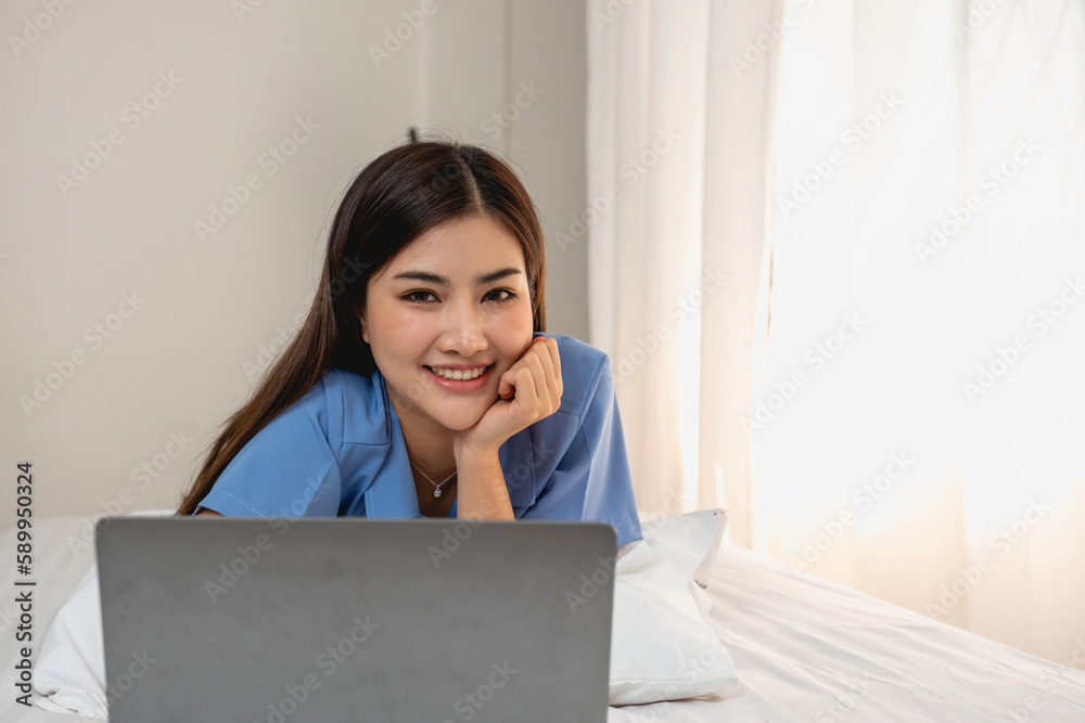 Young Asian woman with beautiful face, long hair, blue shirt lying on bed in white bedroom at home with cup of coffee and video call with laptop talking to relatives on vacation. holiday concept. Young Asian woman with beautiful face, long hair, blue shirt lying on bed in white bedroom at home with cup of coffee and video call with laptop talking to relatives on vacation. holiday concept.