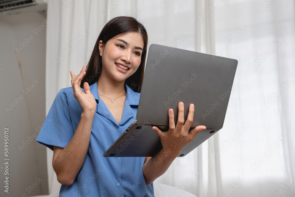 Young Asian woman with beautiful face, long hair in blue shirt sitting on white bedroom bed at home with cup of coffee and video call with laptop talking to relatives on holiday. vacation concept. Young Asian woman with beautiful face, long hair in blue shirt sitting on white bedroom bed at home with cup of coffee and video call with laptop talking to relatives on holiday. vacation concept.