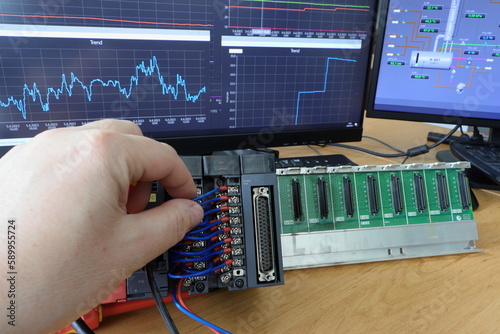 An electrical engineer checks the connection of the PLC automaton. Control system testing. Trends of measured quantities in the background. Production automation. Automatic control system.
