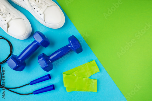 Fitness accessories concept. Photo of elastic band jump rope dumbbells and white sneakers on a two tone green and blue background