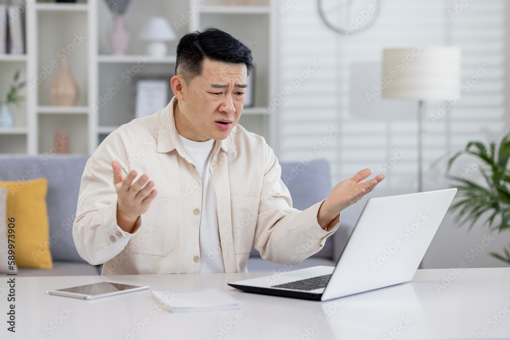 Angry man at home working with laptop, frustrated asian man looking at laptop screen got bad news and unsatisfactory work result, freelancer working inside home office.