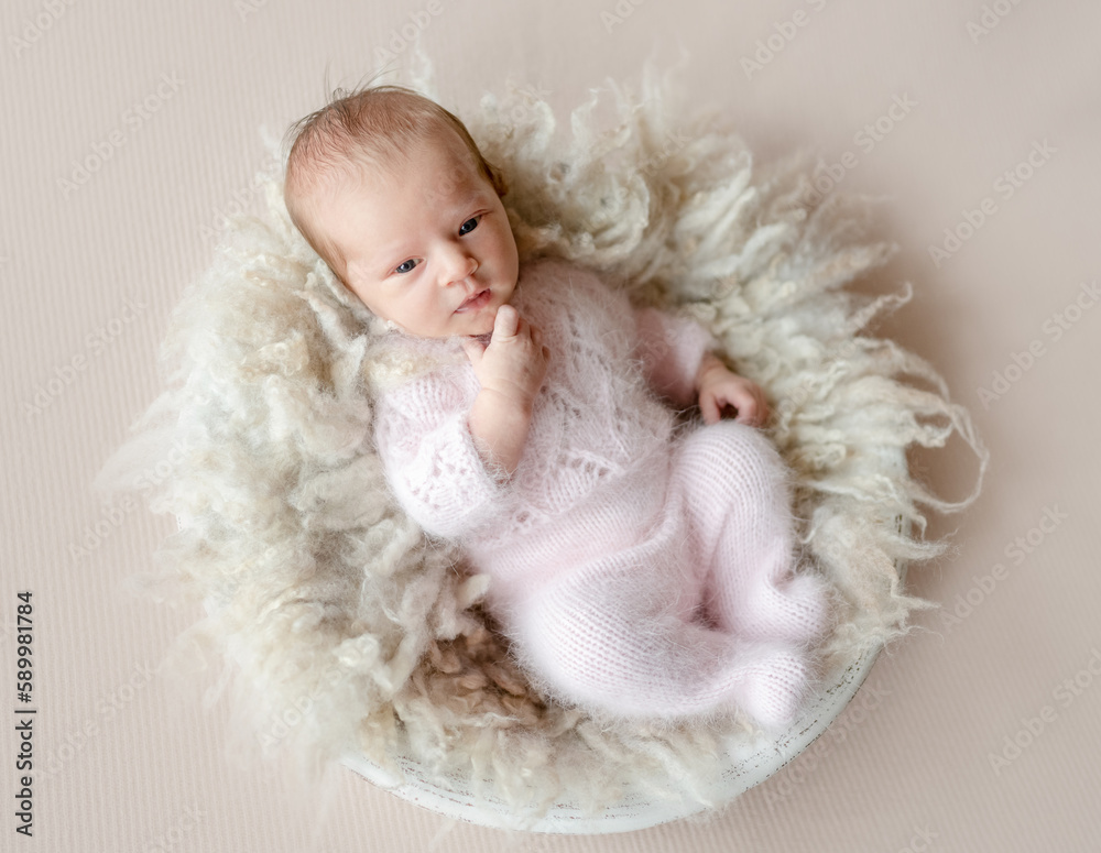 Newborn baby girl wearing knitted costume and looking at camera with thinking face expression. Infant child kid on fur studio portrait Newborn baby girl wearing knitted costume and looking at camera with thinking face expression. Infant child kid on fur studio portrait