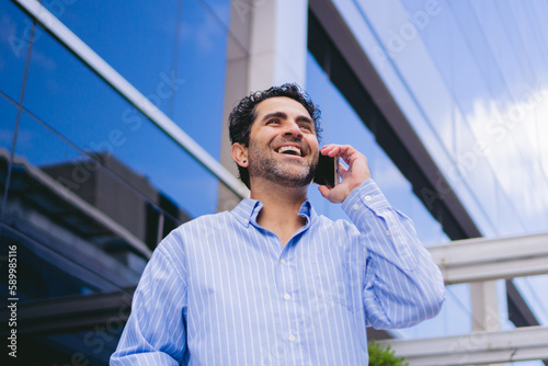 Low angle shot of a middle-aged latin man talking on the phone while laughing in front of a corporate building.