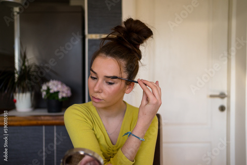 Young teenage woman doing make up in her living room, using eyebrow pencil. 