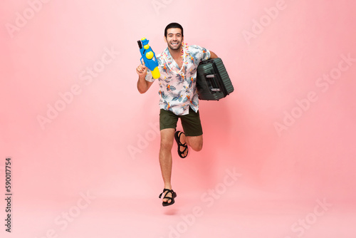 Happy handsome Caucasian tourist man in Thai summer Songkran outfit jumping with water gun and suitcase in pink color studio isolated background