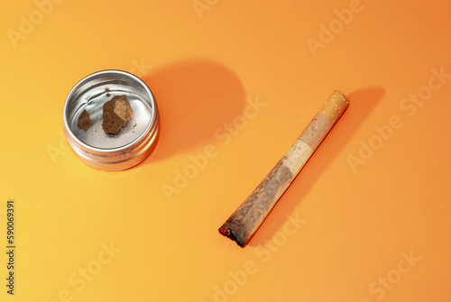 Cigarro porro y pequeño recipiente metálico con trozos de hachís en su interior sobre fondo rugoso de color naranja. photo