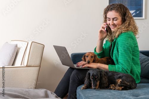 One blond woman wearing green outfit sitting on the couch working on the laptop and two Dachshund dogs 