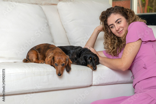 One blond woman wearing pink dress sitting on the floor petting her two Dachshund dogs on the couch in the living room