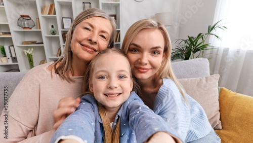 Happy grandmother, mother and granddaughter are sitting on the sofa in the living room, taking selfies, having a video call with relatives, waving. They listen to congratulations on Mother's Day.
