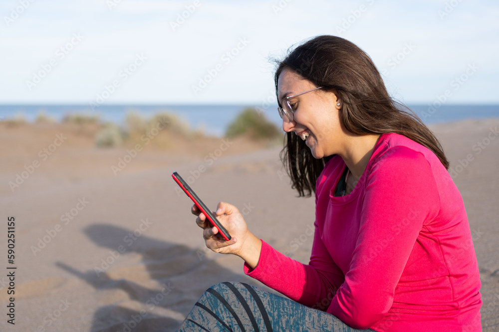 young adult latina brunette woman using a cell phone and smiling on the dunes of a beach in the province of Buenos Aires, Argentina.