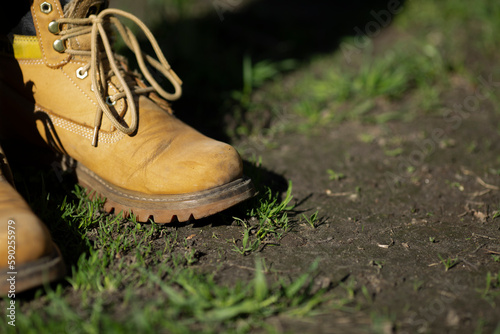 Sand-colored old farm male boots on garden soil. Man stands in garden on lawn with grass breaking through ground. Preparing for spring sowing season. Autumn. Farmer. Worker in safety shoes. Copyspace