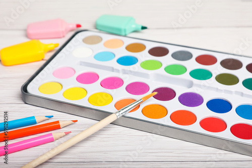 Watercolor palette with brush, colorful pencils and markers on white wooden table, closeup
